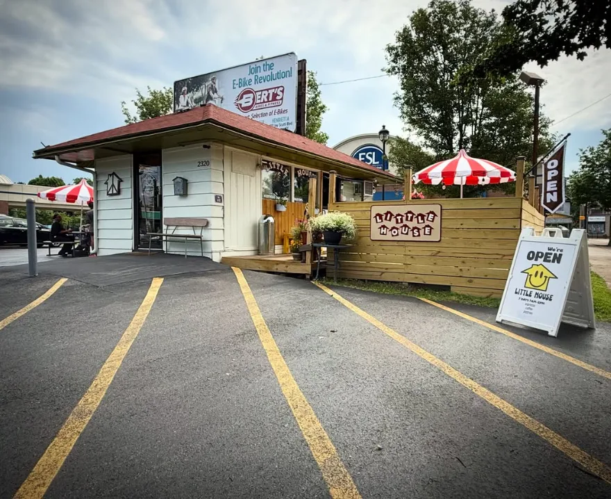The shop from the parking lot: a small white building with a red roof, a red-and-white umbrella, a fenced patio and an Open sign