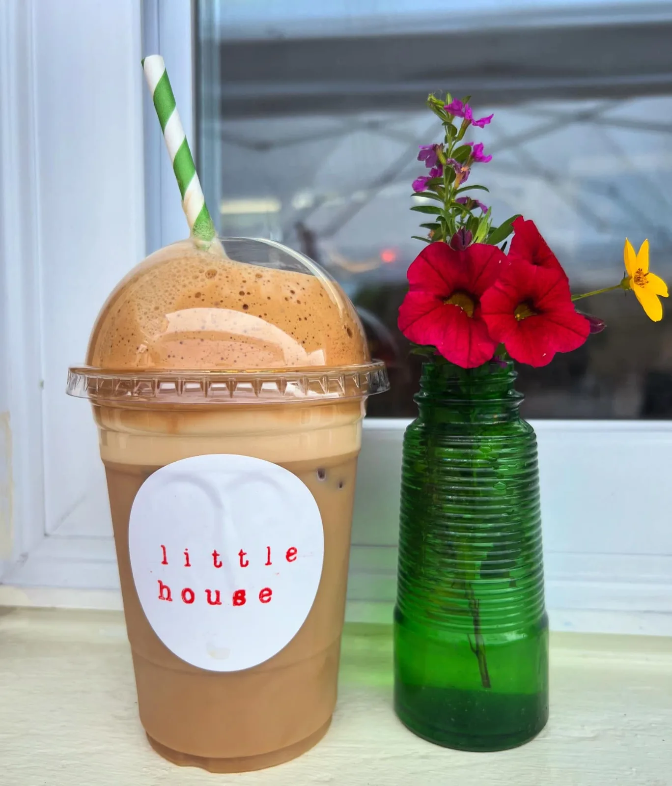 An iced latte with a Little House sticker and a striped straw, beside a small vase of flowers