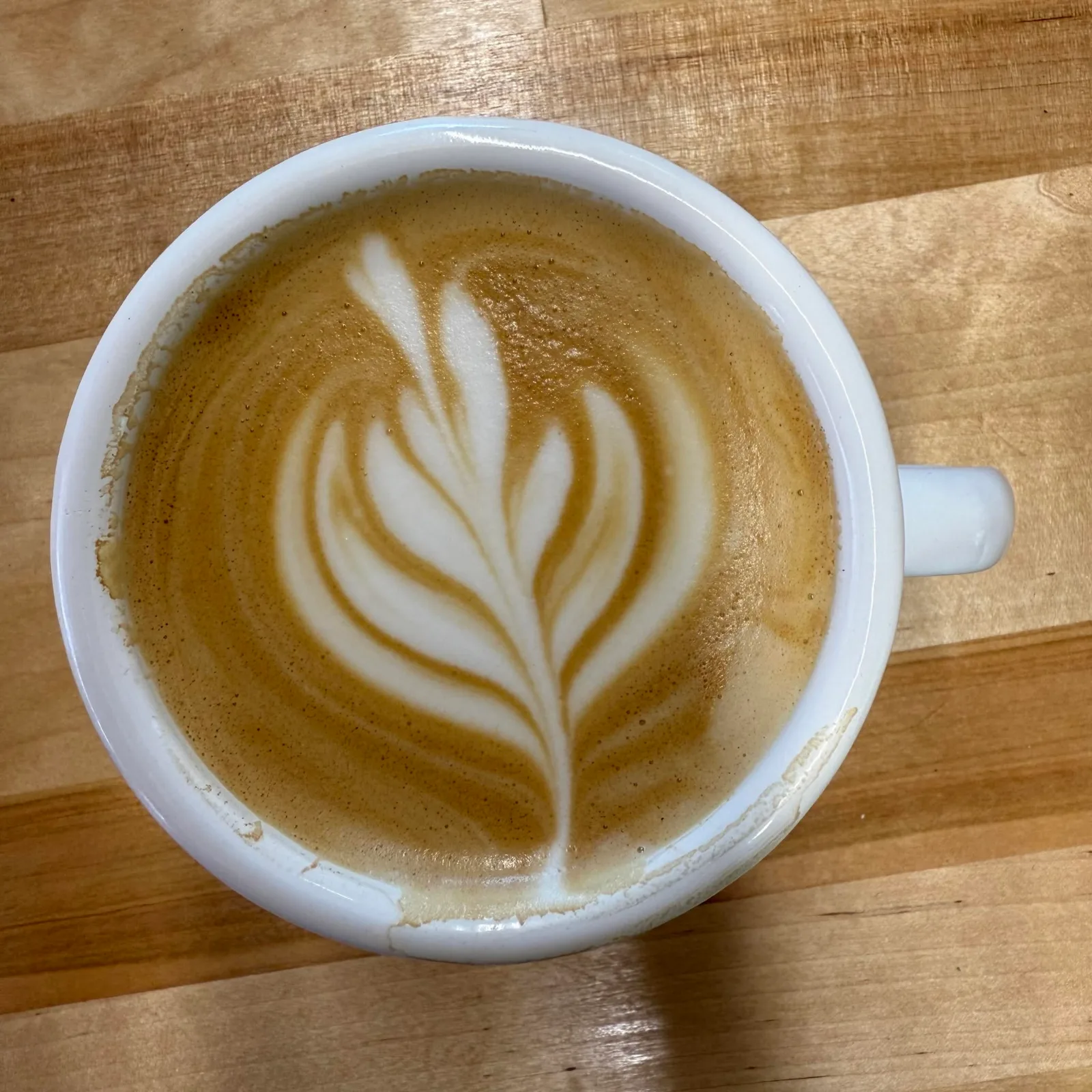 A latte with a leaf poured in the foam, in a white cup on a wooden counter