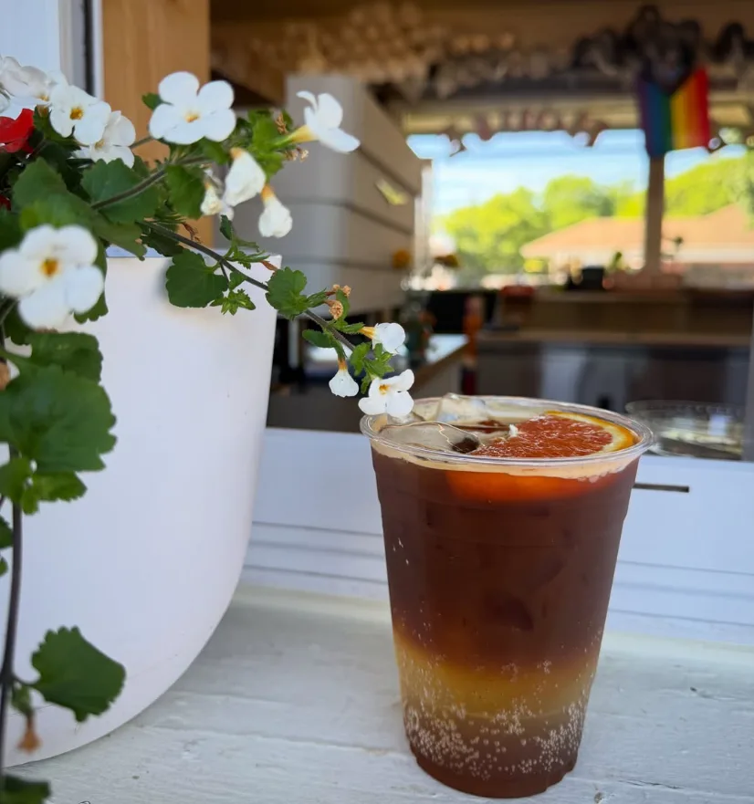An espresso tonic over ice with an orange slice, on a windowsill beside white flowers