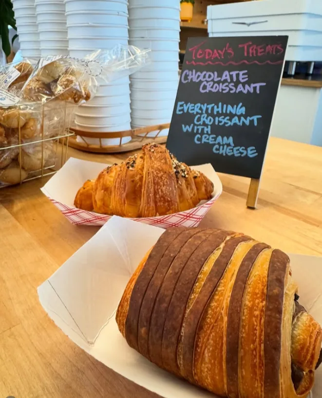 A chocolate croissant and an everything croissant, with the day’s treats written on a chalkboard behind them