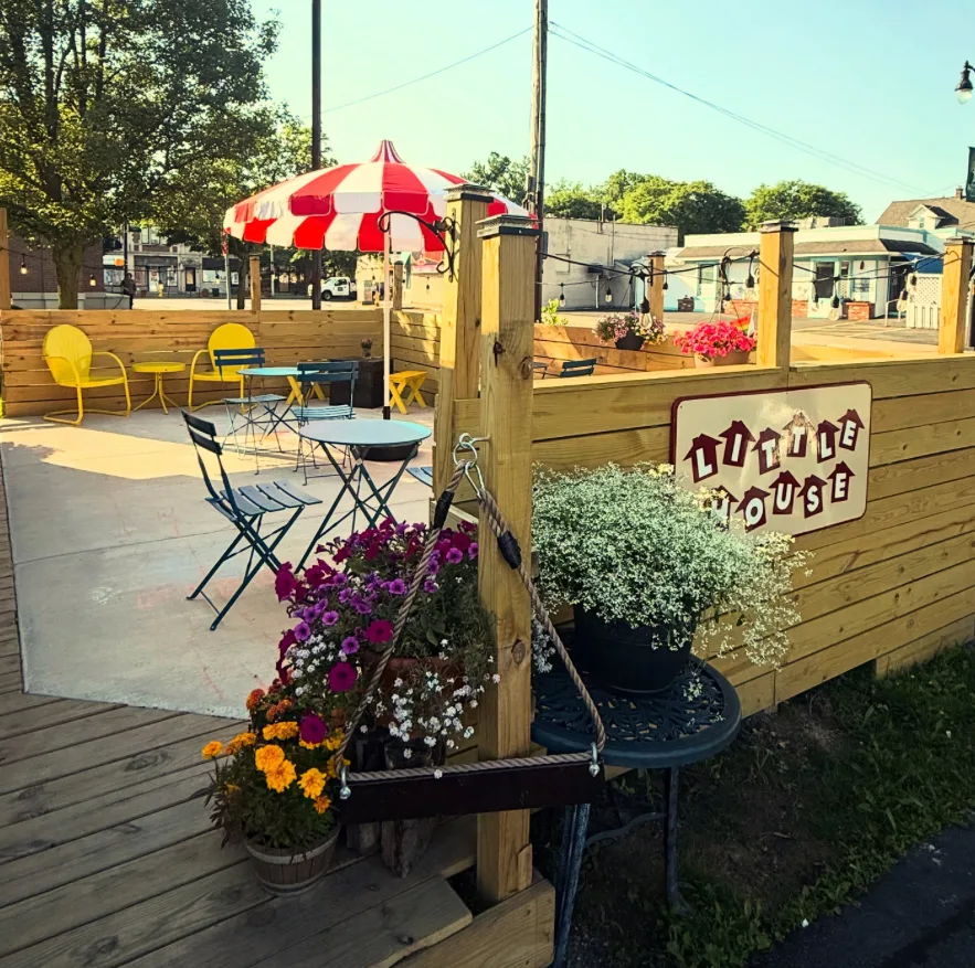 The patio beside the shop: yellow chairs, bistro tables and a red-and-white umbrella behind a wooden fence with the Little House sign